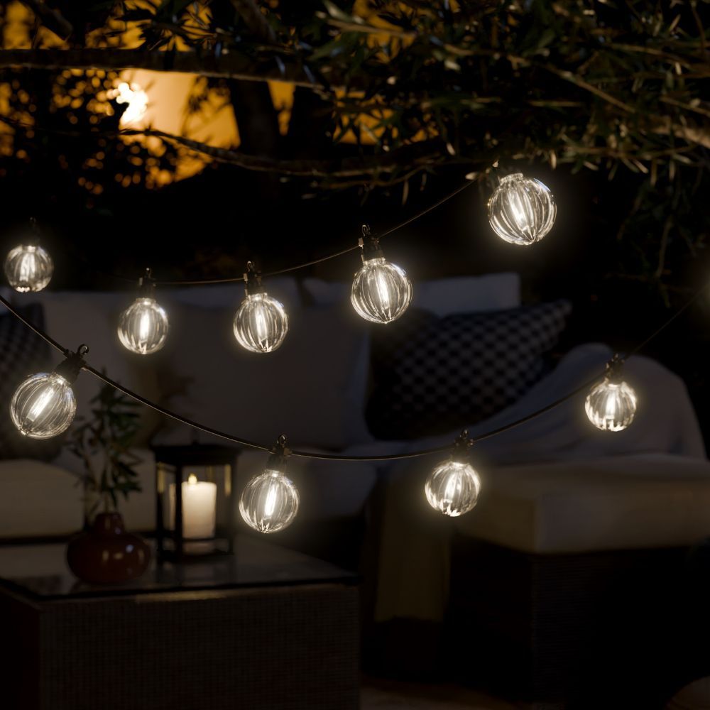 Lyskæde med stribede LED-kugler over loungesofa med puder og lanterne i haven om aftenen