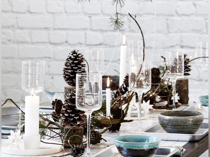 Nordisk juleborddækning med hvide stearinlys, høje glas, grankogler, grene og en diskret lyskæde i messing foran en hvid murstensvæg.