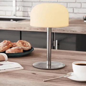 Tændt grå bordlampe med varmt lys på køkkenbord med kaffe, kanelsnegle og magasin